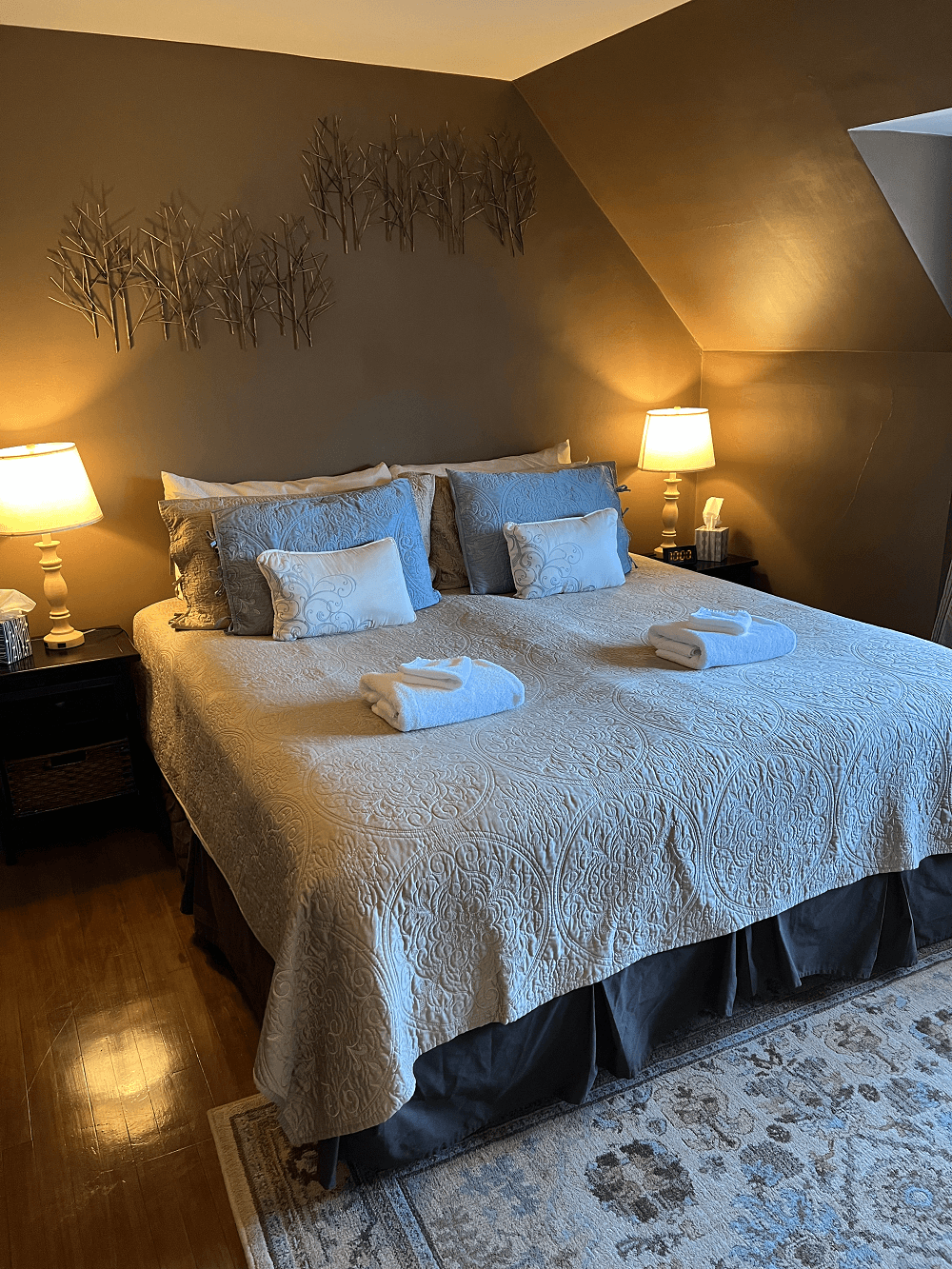 A cozy bedroom with chocolate brown walls and a sloped ceiling featuring a king bed with beige quilted linens, blue accent pillows, and folded white towels. Warm bedside lamps flank the bed, and a patterned area rug rests on the hardwood floor.
