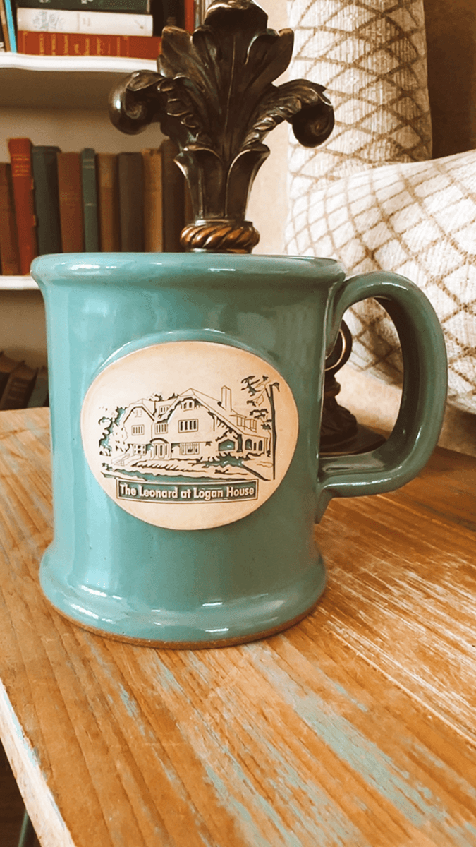 A turquoise mug featuring an emblem of "The Leonard at Logan House" on a wooden table.