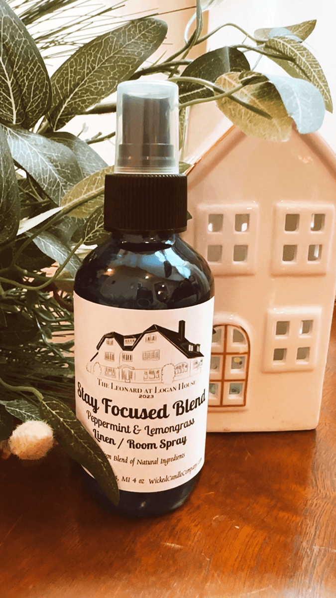 A bottle of peppermint and lemongrass room spray is placed next to a small ceramic house and greenery on a wooden surface.