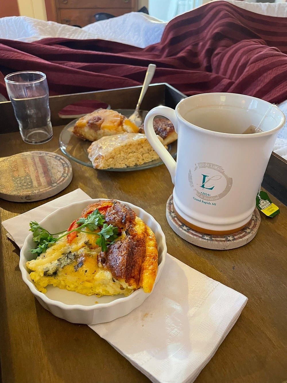 A breakfast tray featuring an omelet, pastries, a glass of water, and a cup of tea, set against a cozy backdrop.