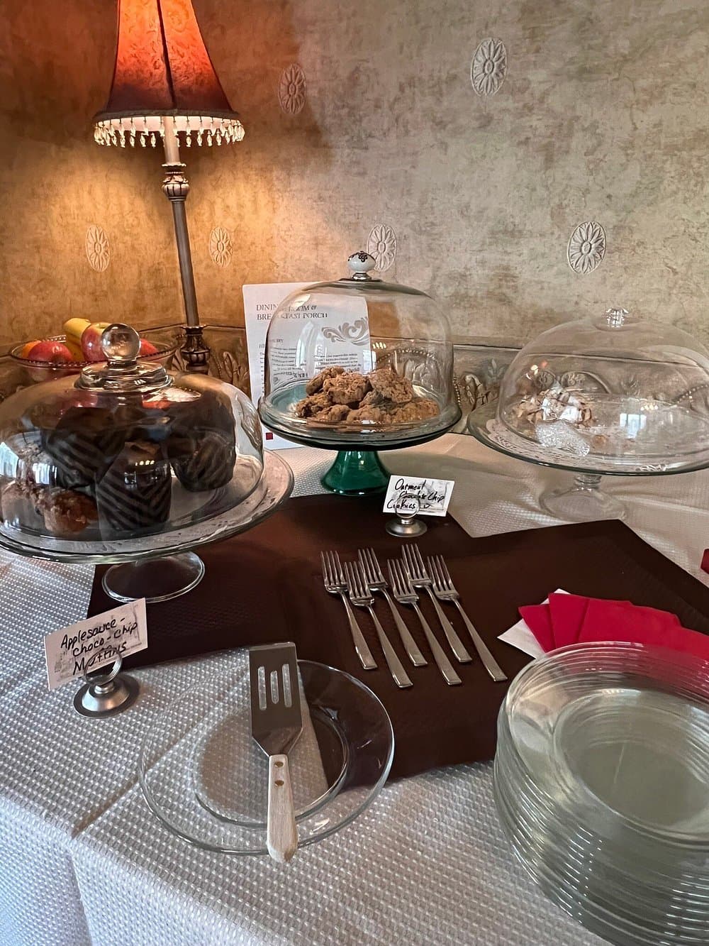A dessert table features various cookies and muffins under glass cloches, accompanied by utensils and plates.