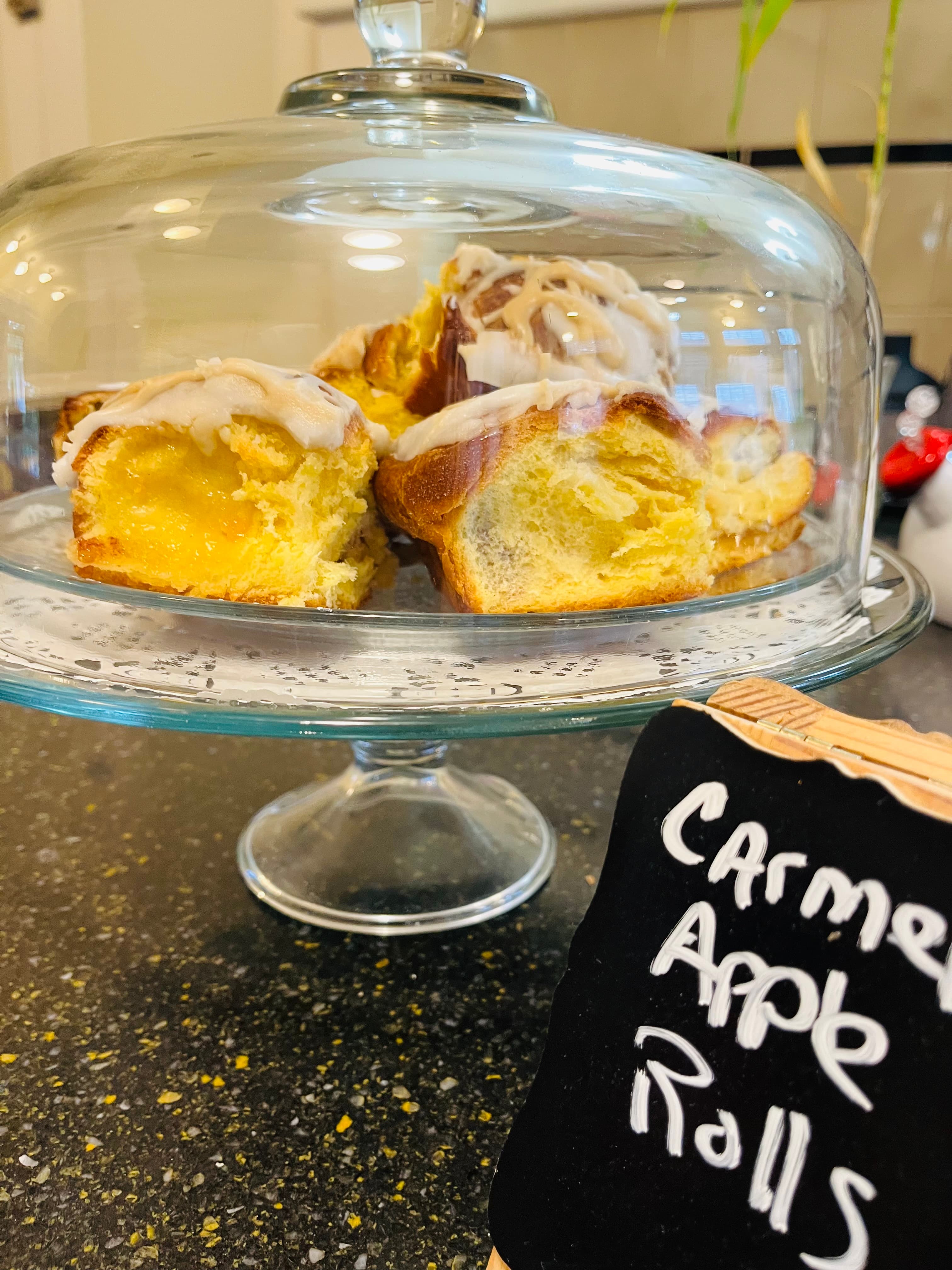 A glass cake dome displays caramel apple rolls, with a black chalkboard sign labeling them.
