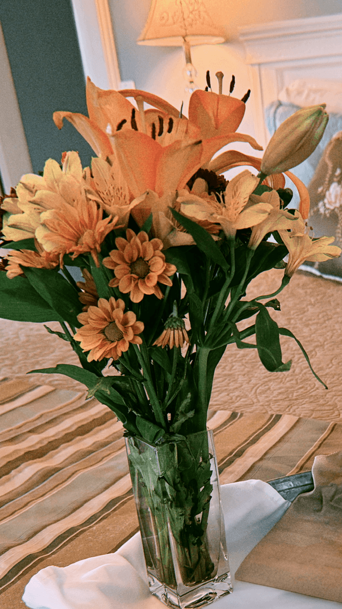 A bouquet of orange flowers in a tall glass vase sits on a patterned tablecloth.
