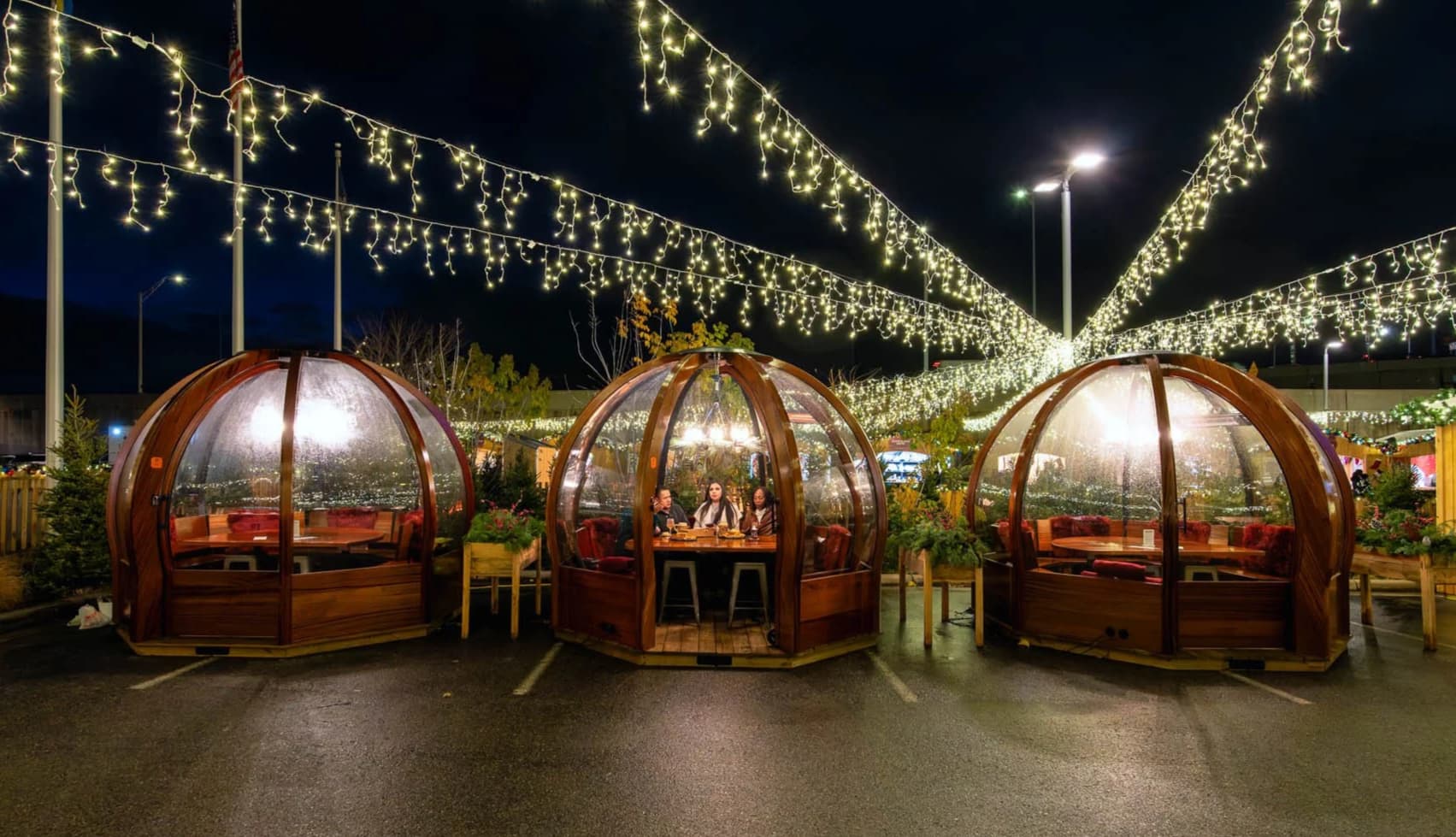 Dining igloos lit with string lights at night. Two wooden igloos house people dining, surrounded by greenery, creating a cozy, festive atmosphere. Dining igloos lit with string lights at night. Two wooden igloos house people dining, surrounded by greenery, creating a cozy, festive atmosphere.